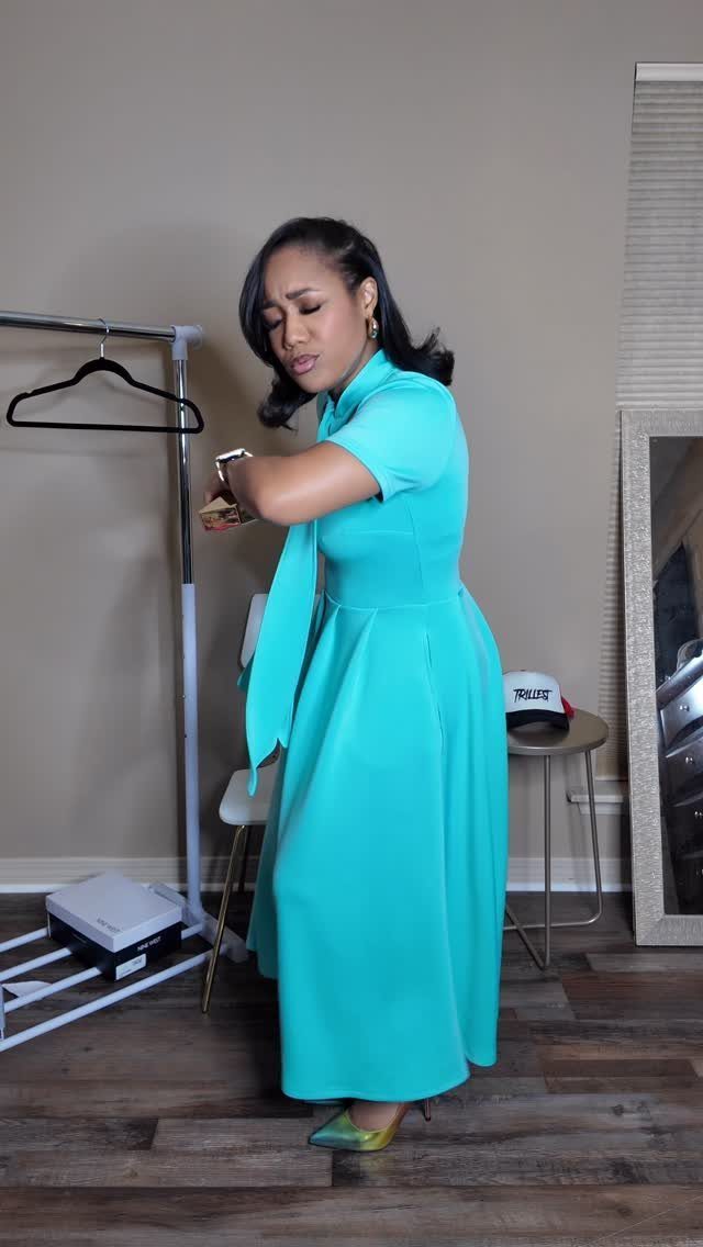 Woman in a turquoise midi dress adjusting her watch in a room with a clothing rack.