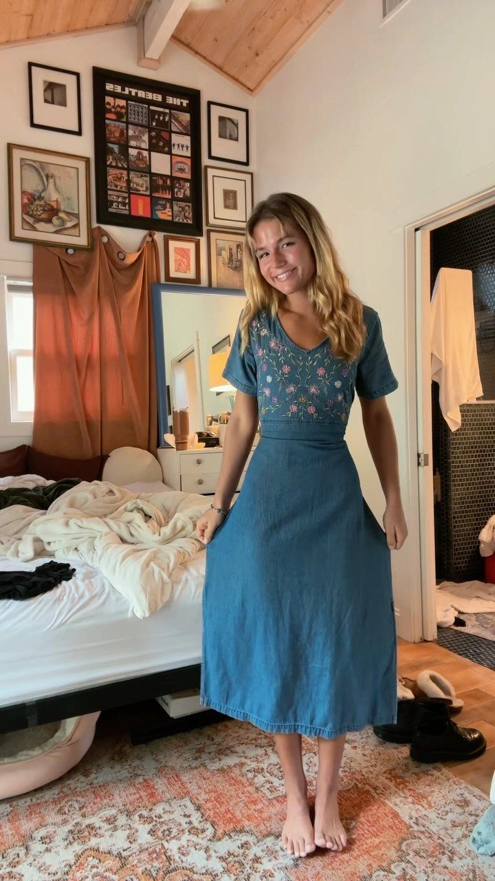 A smiling young woman standing in a bedroom wearing a blue floral embroidered denim midi dress.