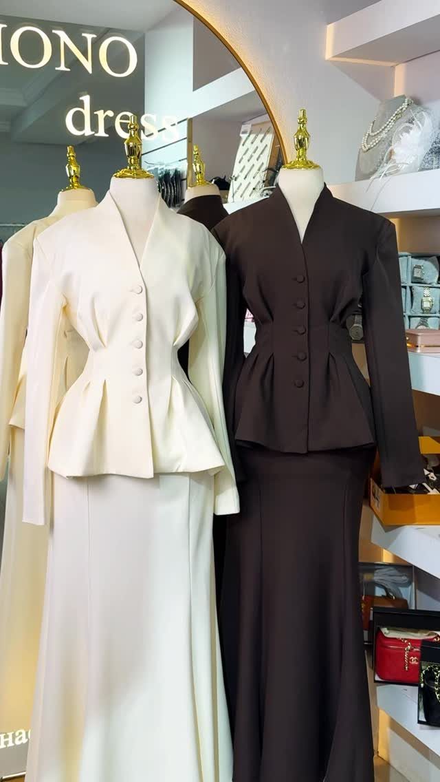 Two mannequins wearing formal cream and brown peplum jackets with matching long skirts in a boutique.
