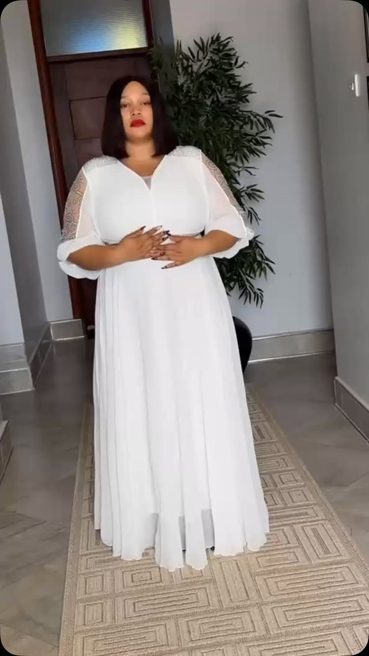A woman posing in a long, elegant white dress with sheer embellished sleeves.