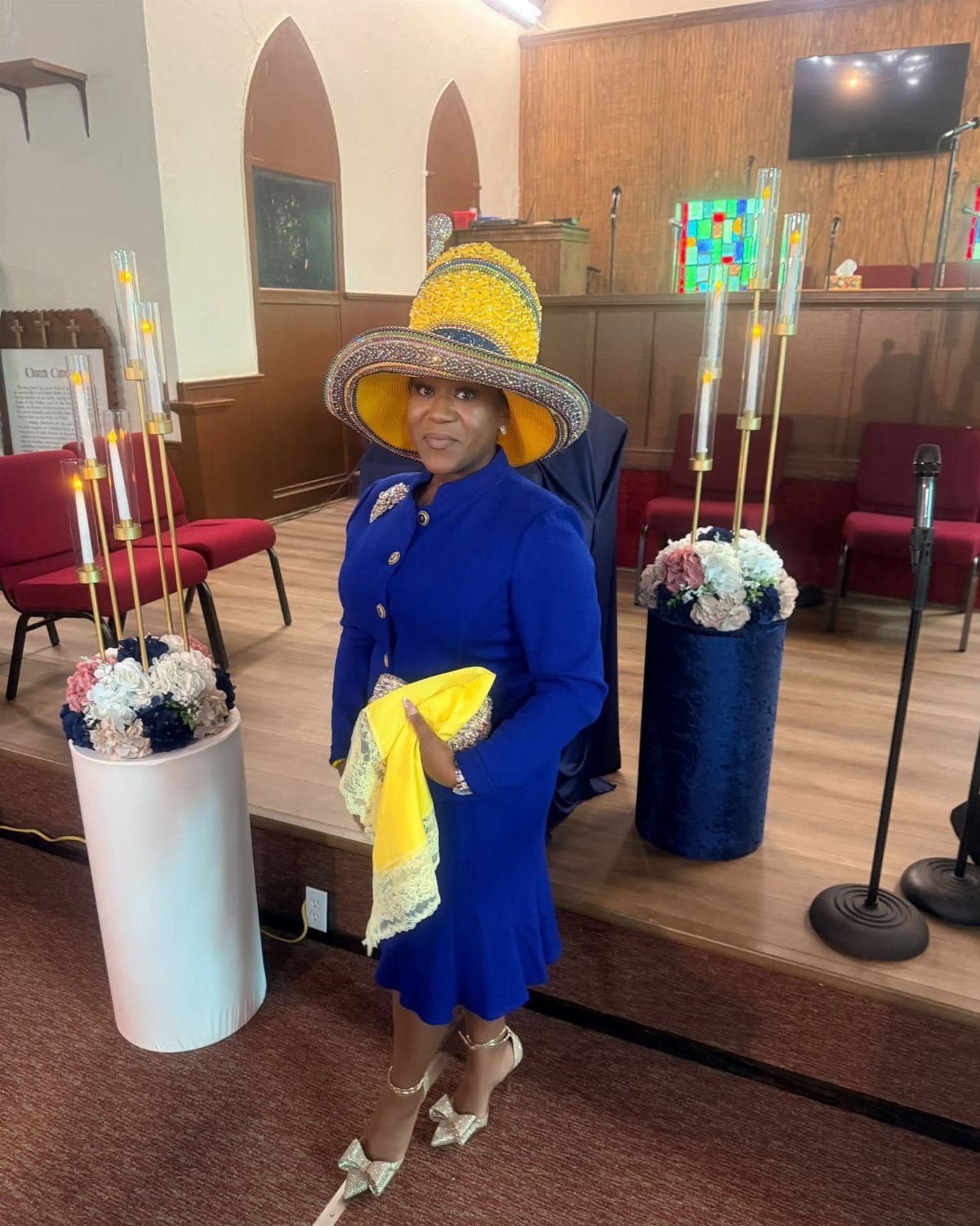Woman in a royal blue church suit and large yellow hat posing indoors.