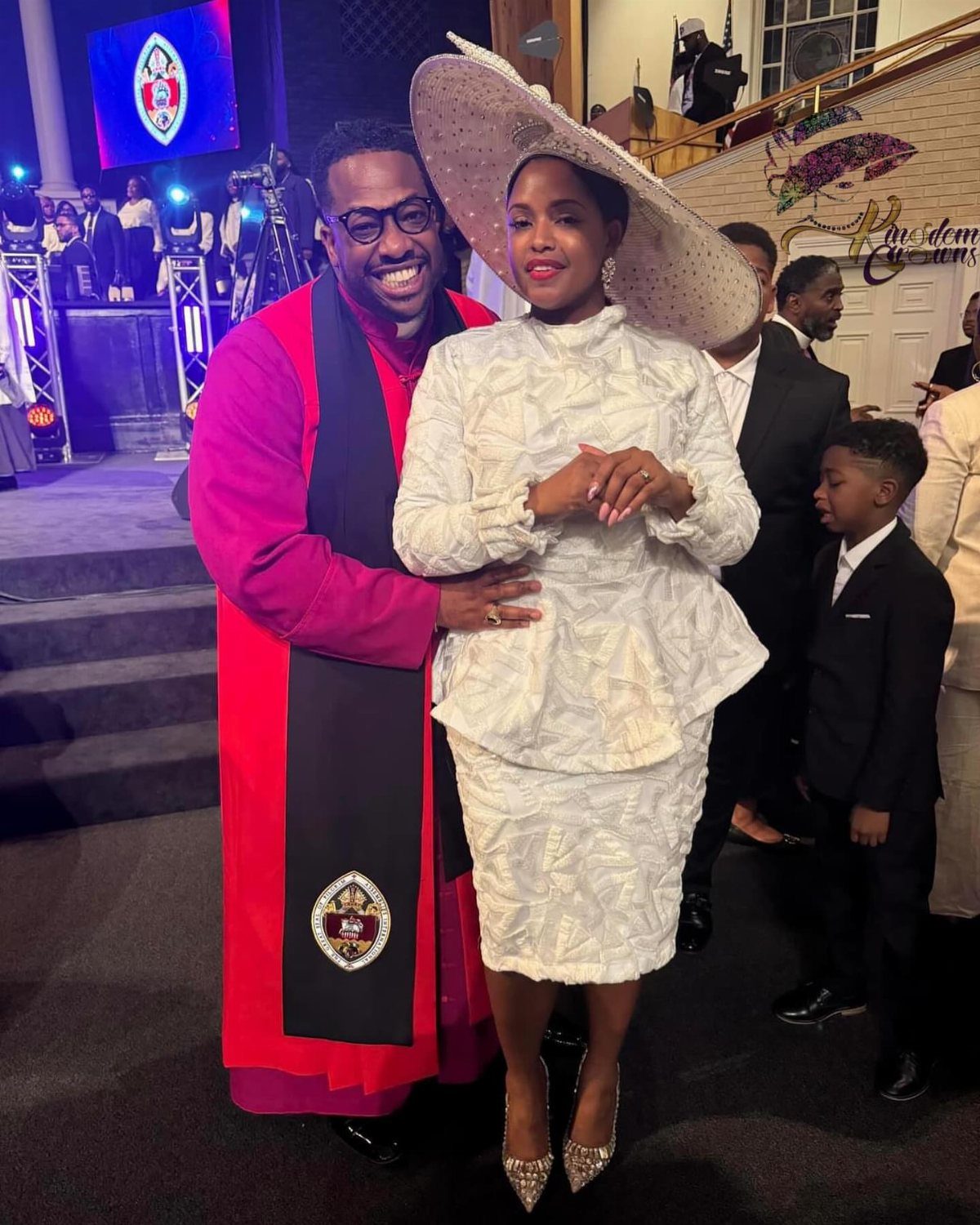Woman in a textured white peplum suit and large hat standing next to a man in clerical robes.