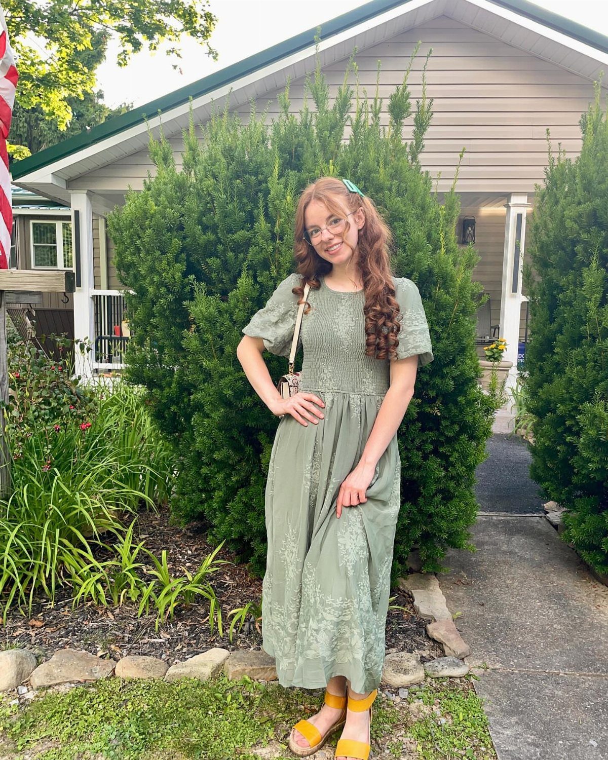 Young woman posing in a sage green maxi dress and yellow sandals in front of bushes.