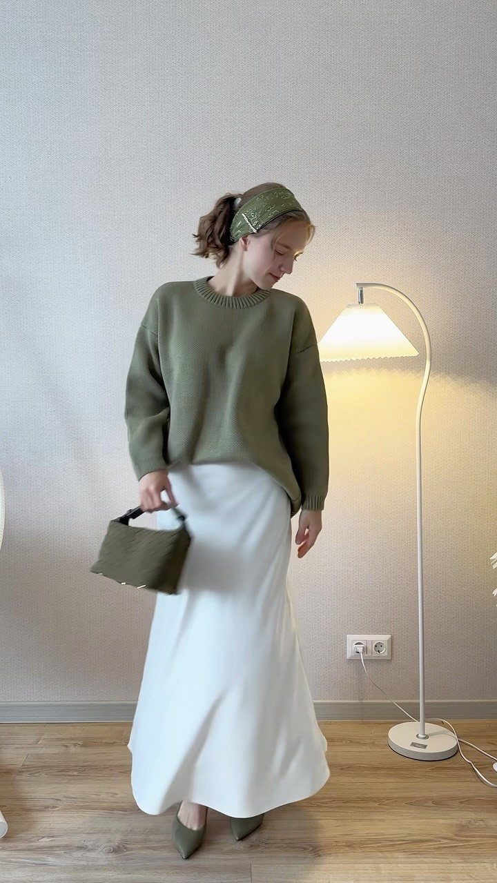 A woman posing in an olive green sweater, long white skirt, and matching green accessories.