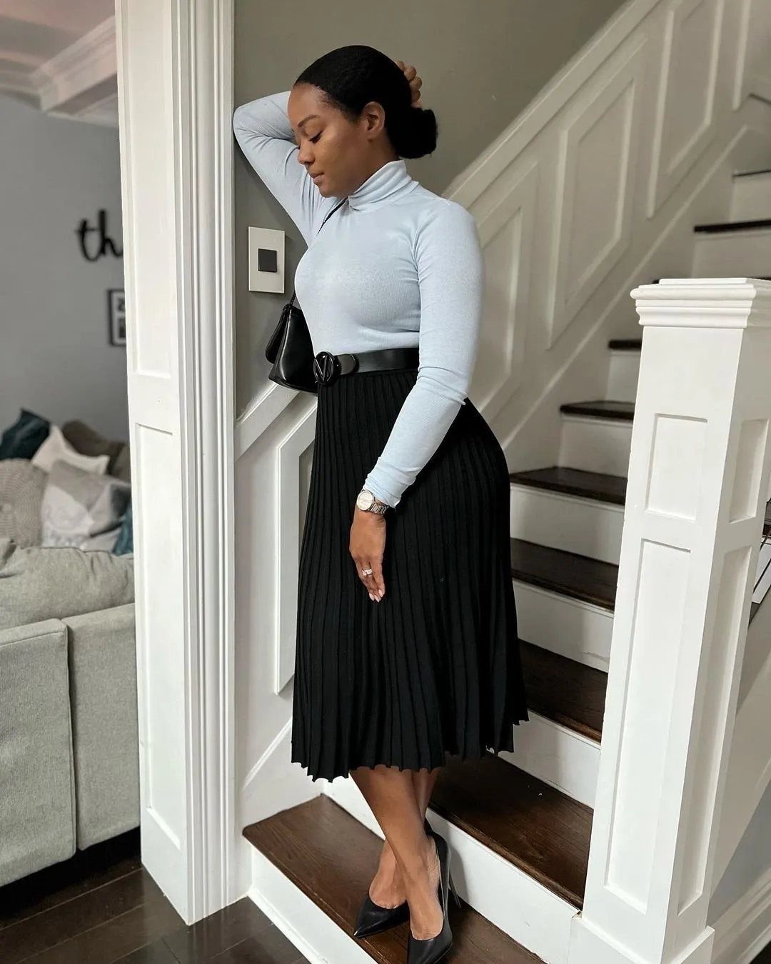 A woman in a light blue turtleneck and black pleated skirt posing on stairs.