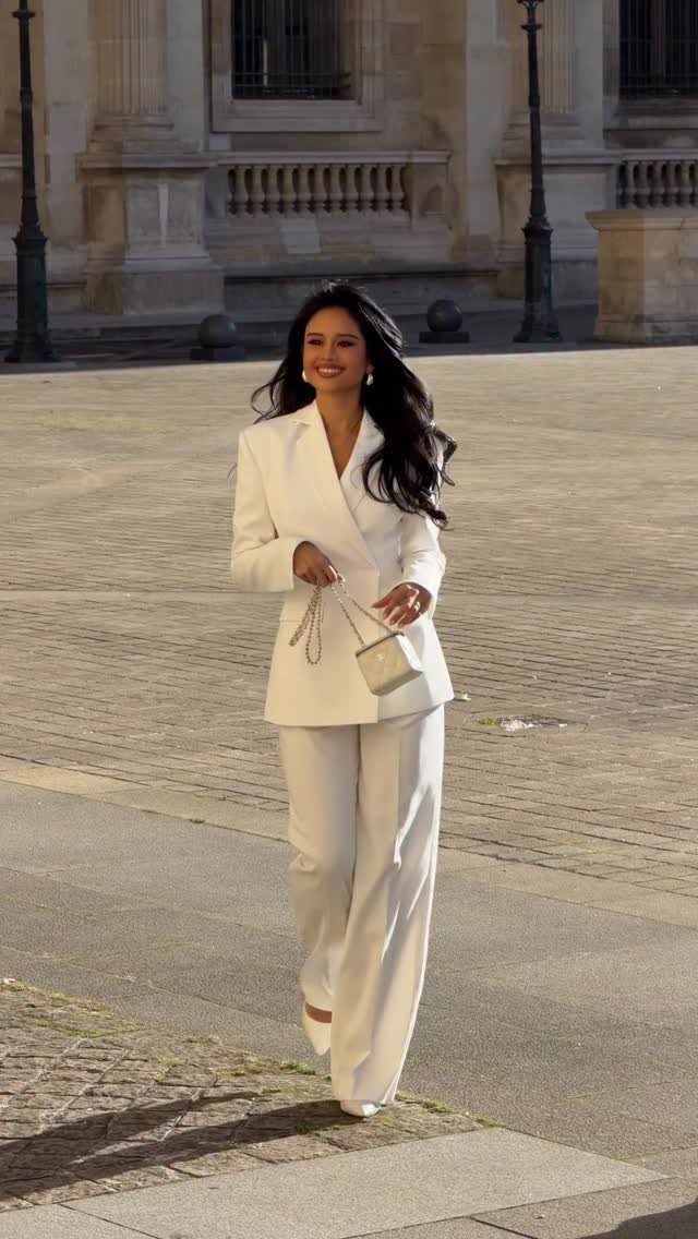 A woman smiling while walking in a white tailored pantsuit and carrying a small white purse.