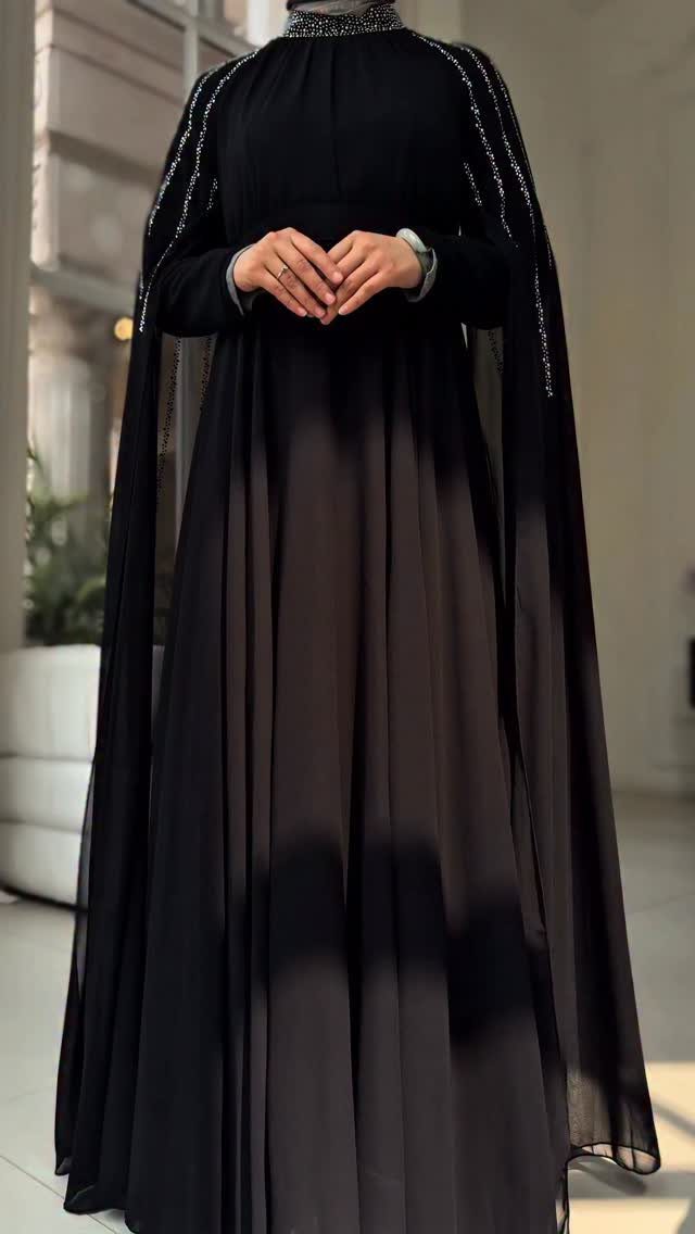A person wearing a long-sleeved black dress with flowing chiffon layers and sparkling shoulder details.