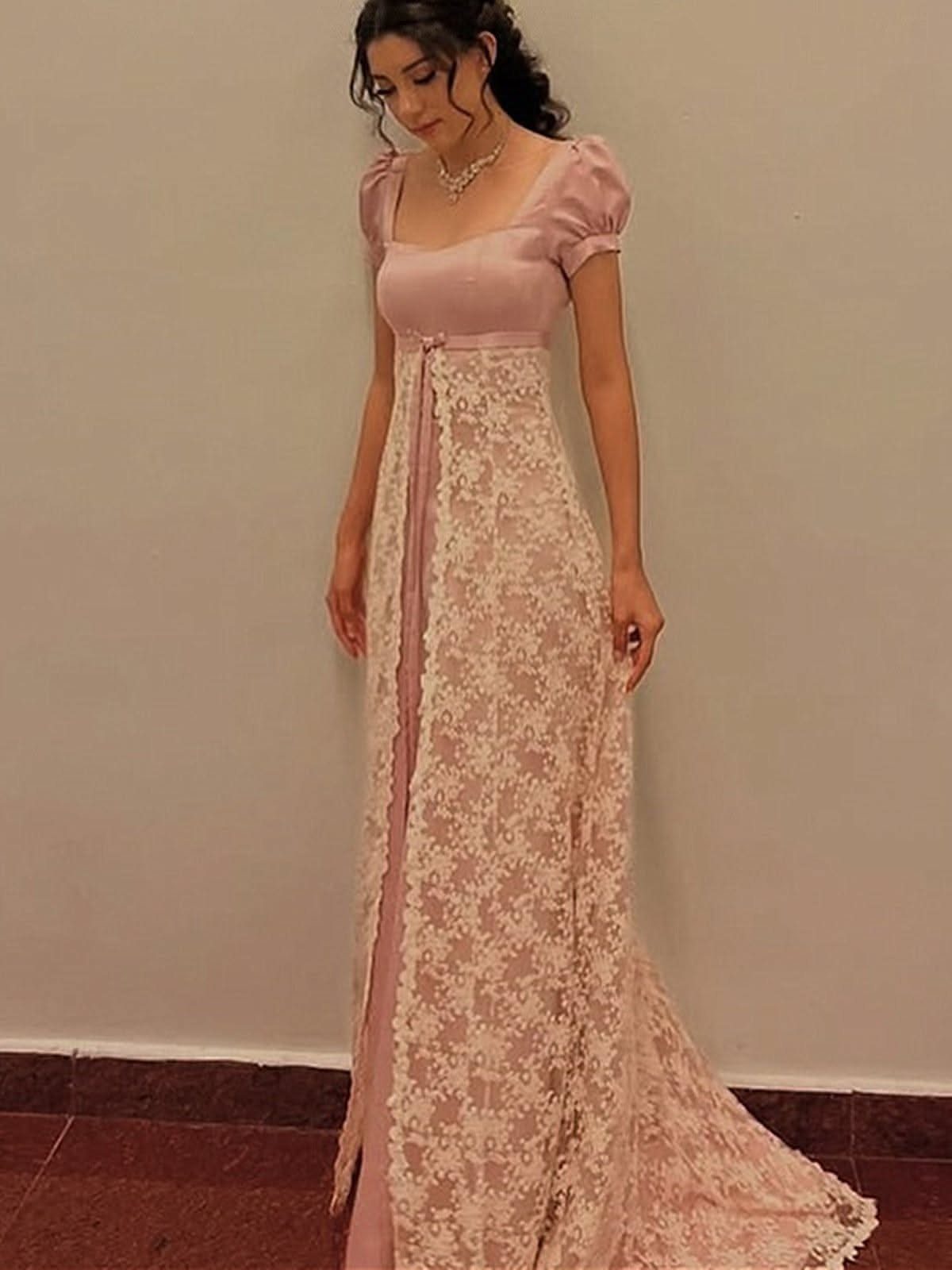 A woman posing in a long, pink empire waist lace gown with a satin bodice and puffed sleeves.