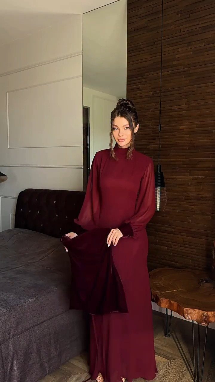 Woman in a long-sleeved burgundy dress with a high neckline standing in a room.