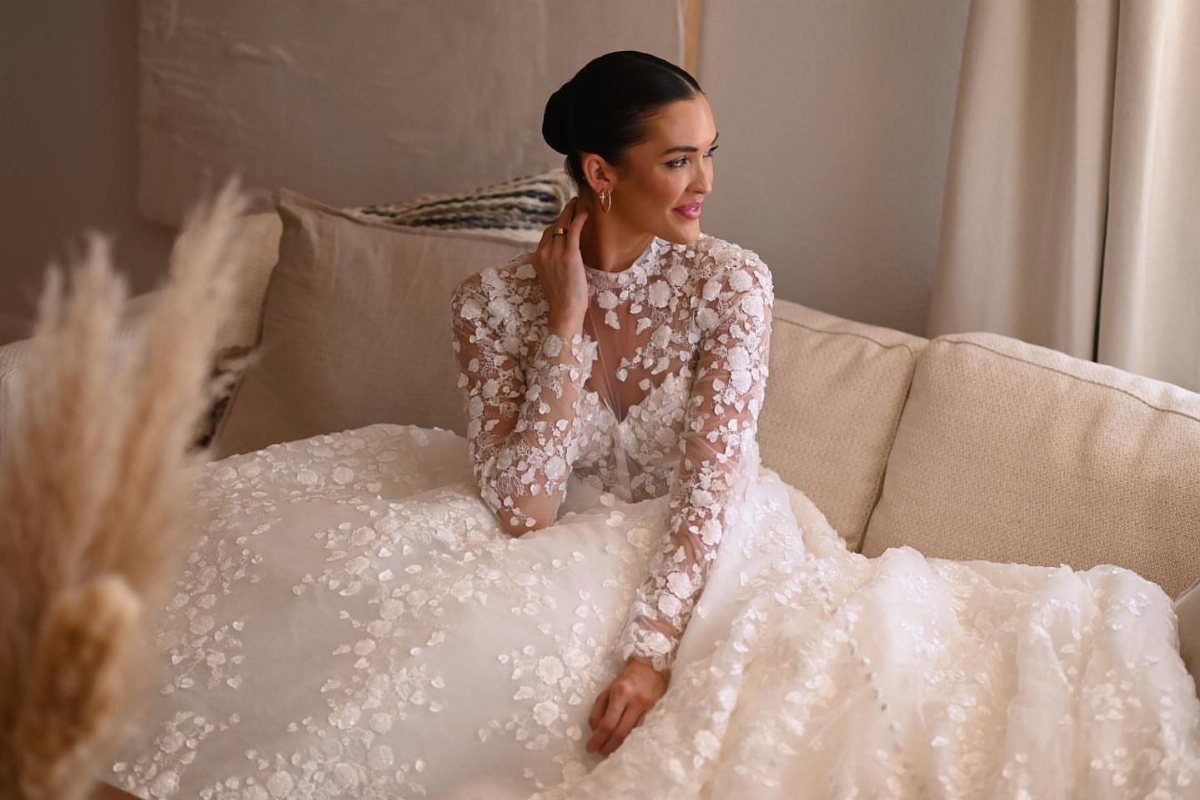 Woman in a white long-sleeved wedding dress with floral illusion lace sitting on a sofa.