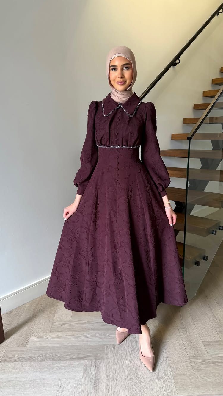 A woman in a long-sleeved purple dress and hijab standing near a staircase.