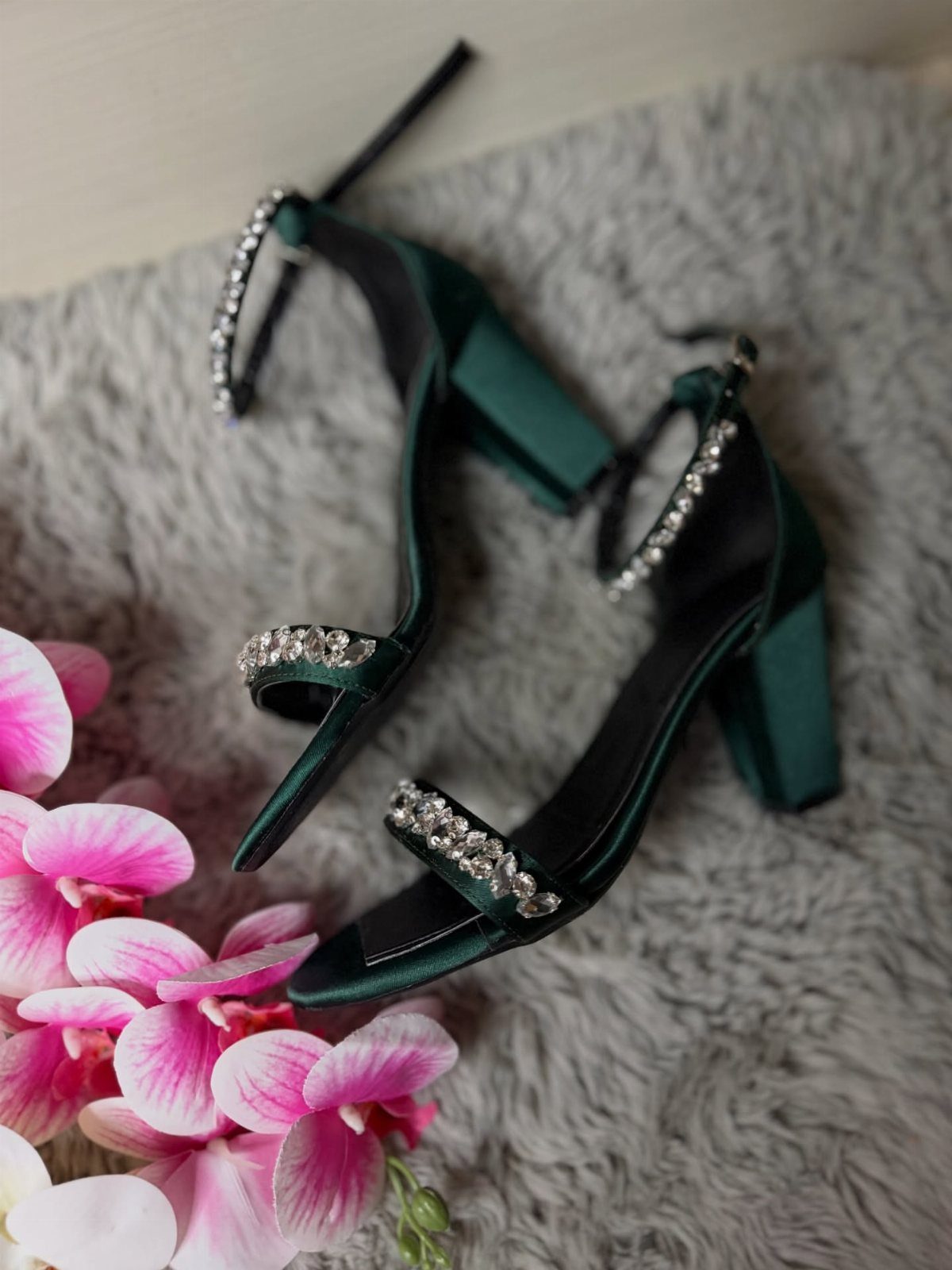 Emerald green satin high heels with rhinestone straps resting on grey fabric near pink orchids.