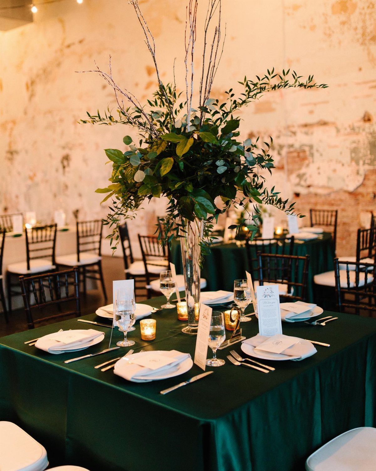 A wedding table set with emerald green linen, white plates, and a tall green foliage centerpiece.