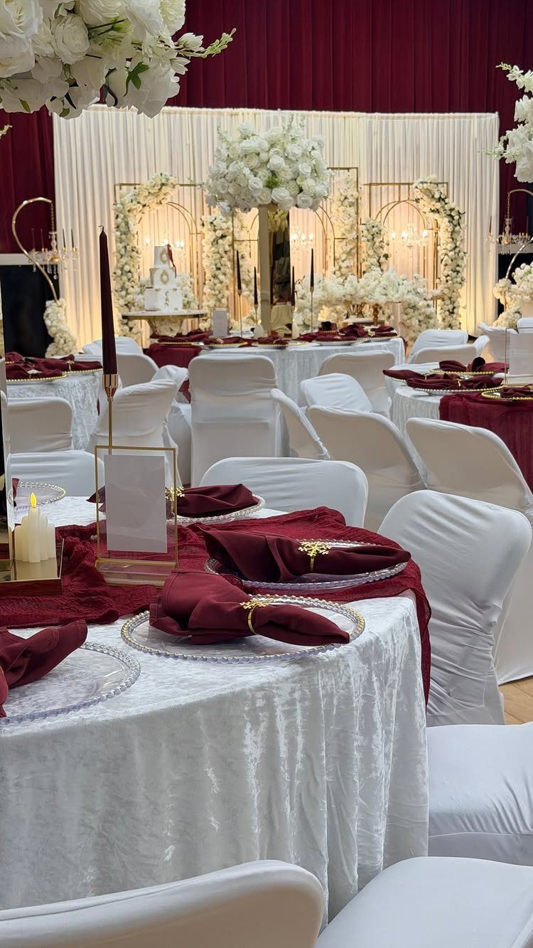 Wedding reception tables with white linens, burgundy napkins, and gold accents with white floral decor.