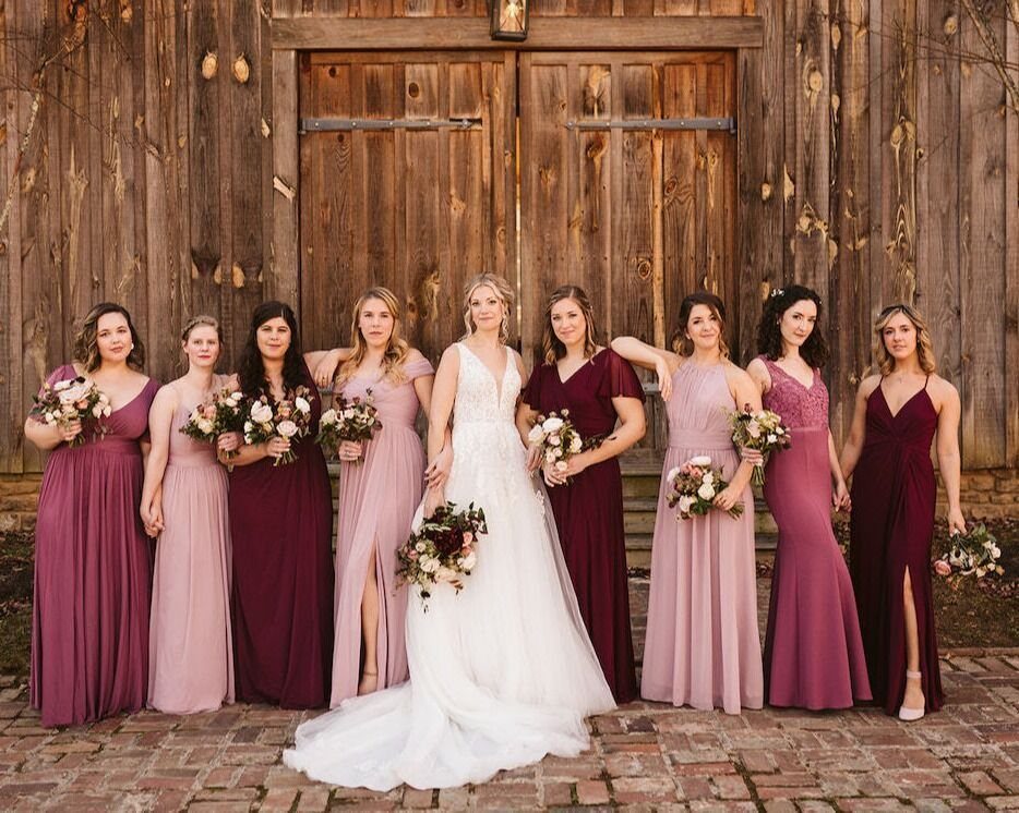 Bridesmaids in dusty rose and plum dresses standing with a bride in front of a barn.