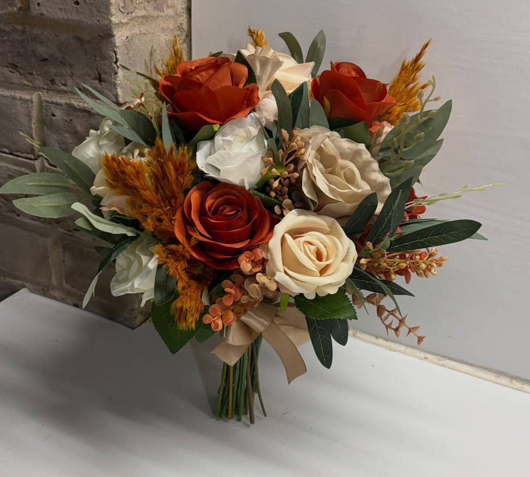 A wedding bouquet with burnt orange, cream, and mustard flowers and green leaves.