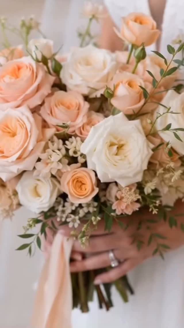Peach and cream roses with green foliage held in a bridal bouquet by a person.