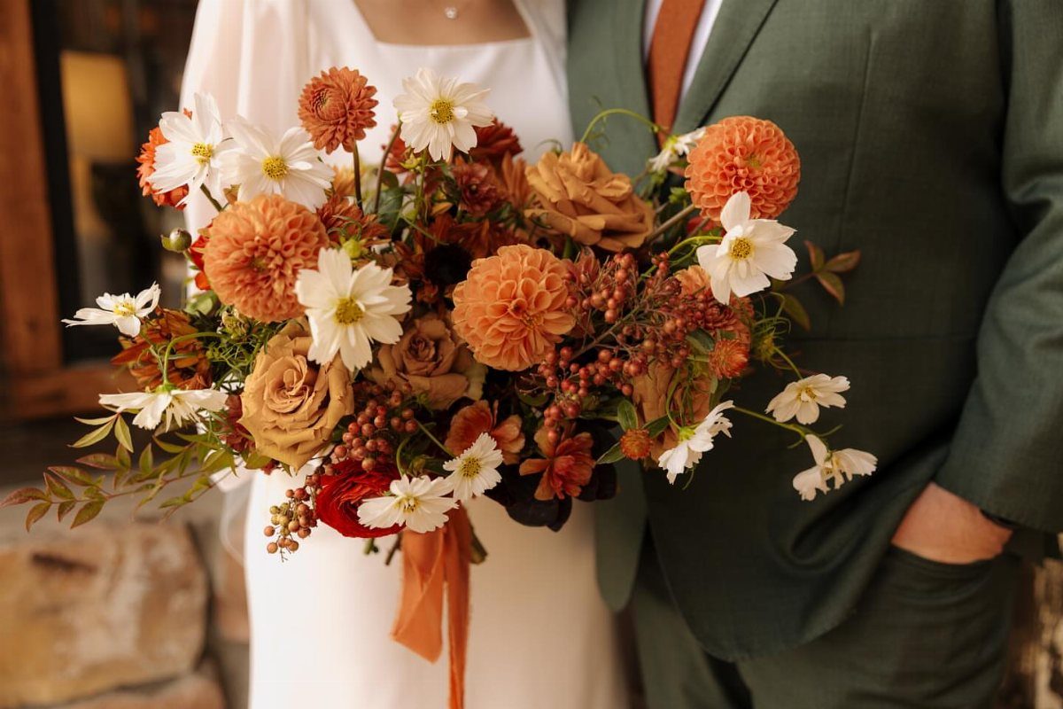 A large floral bouquet with orange dahlias, white flowers, and reddish berries held by a couple.