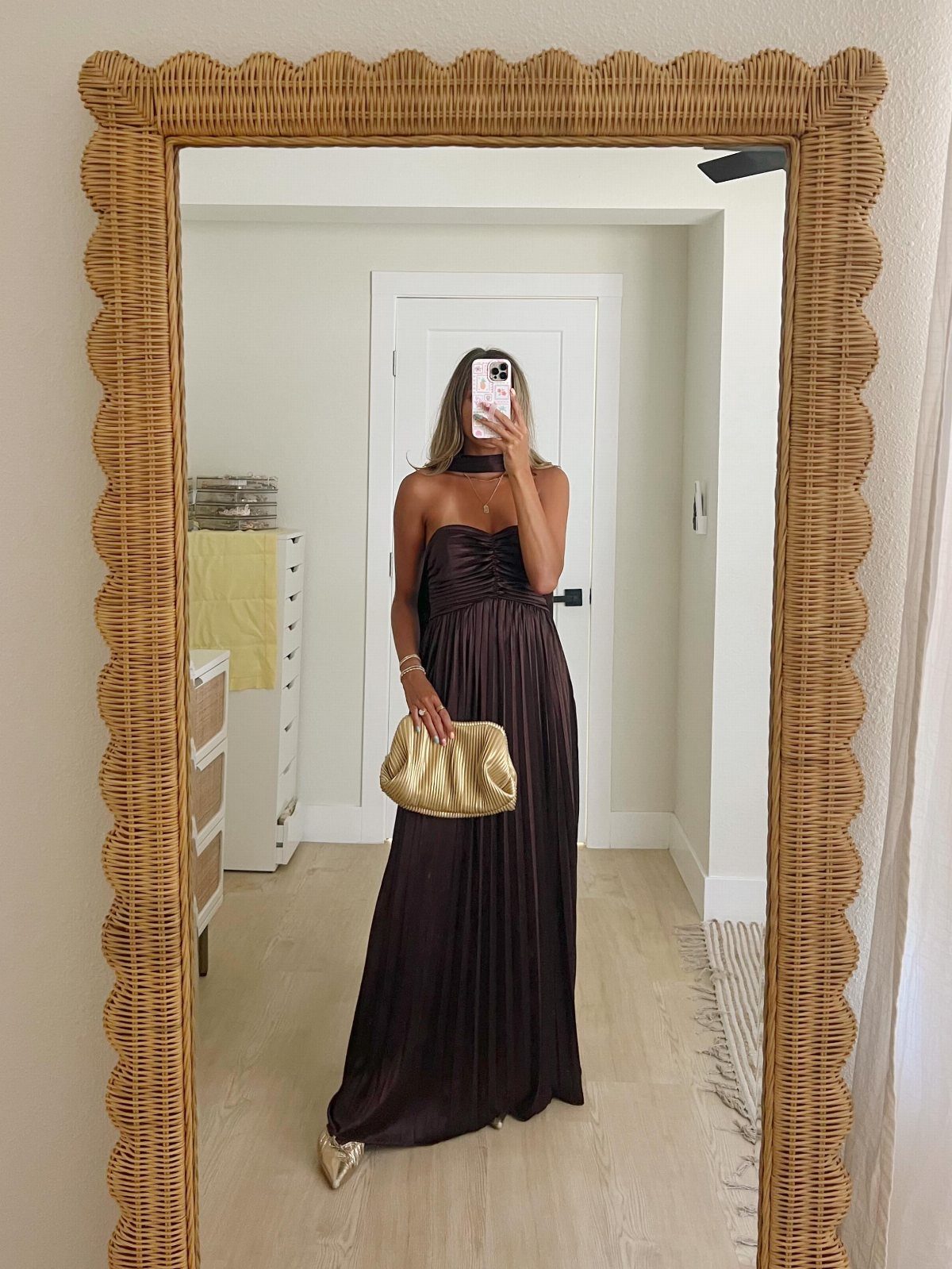 Woman in a long dark brown dress holding a gold clutch in a mirror selfie.