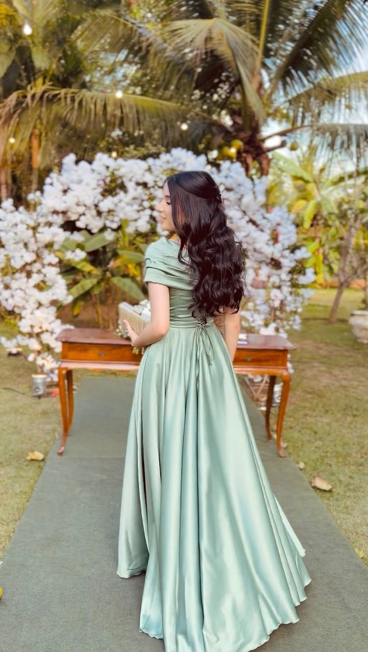 A woman in a long sage green satin dress standing outdoors near white flowers and palm trees.