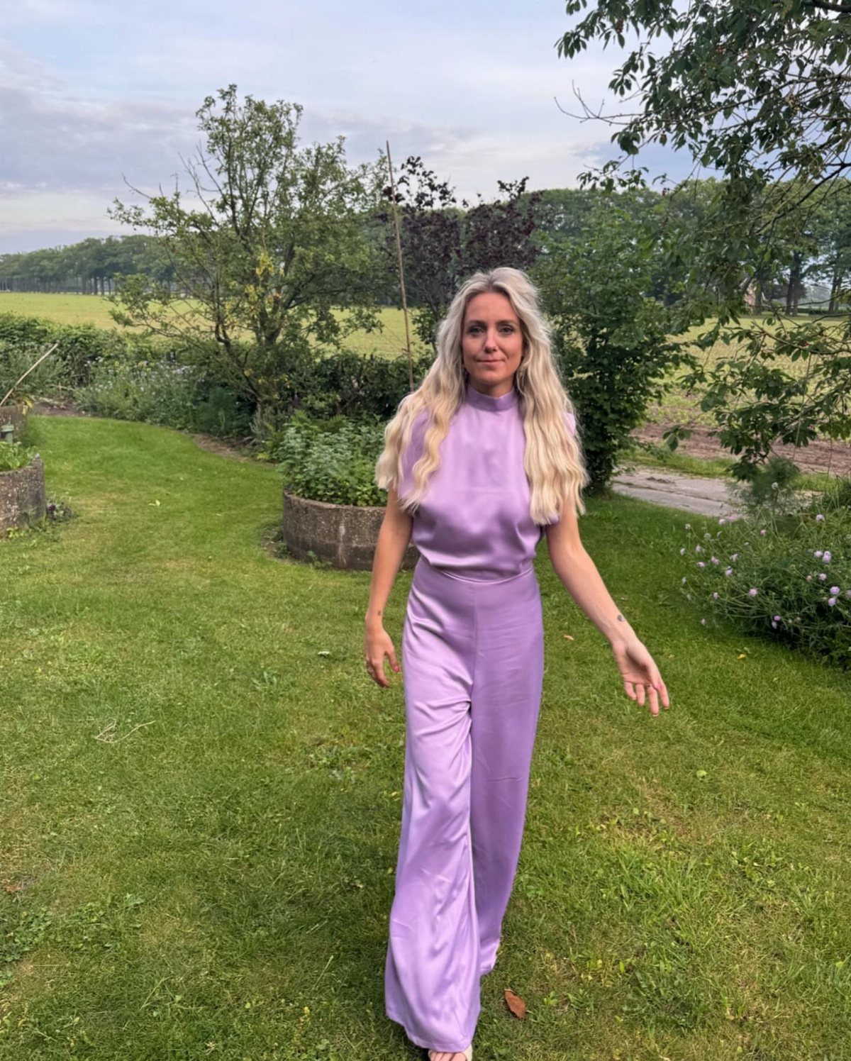 A woman wearing a lilac sleeveless wide-leg jumpsuit standing in a lush green garden.