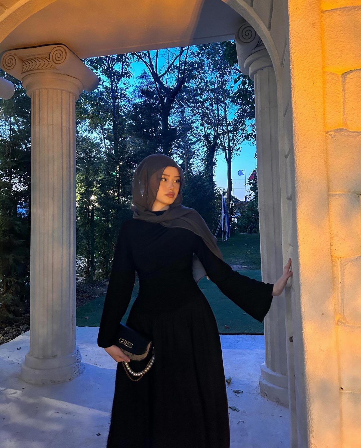 A woman in a black formal dress and grey hijab posing under a white archway.