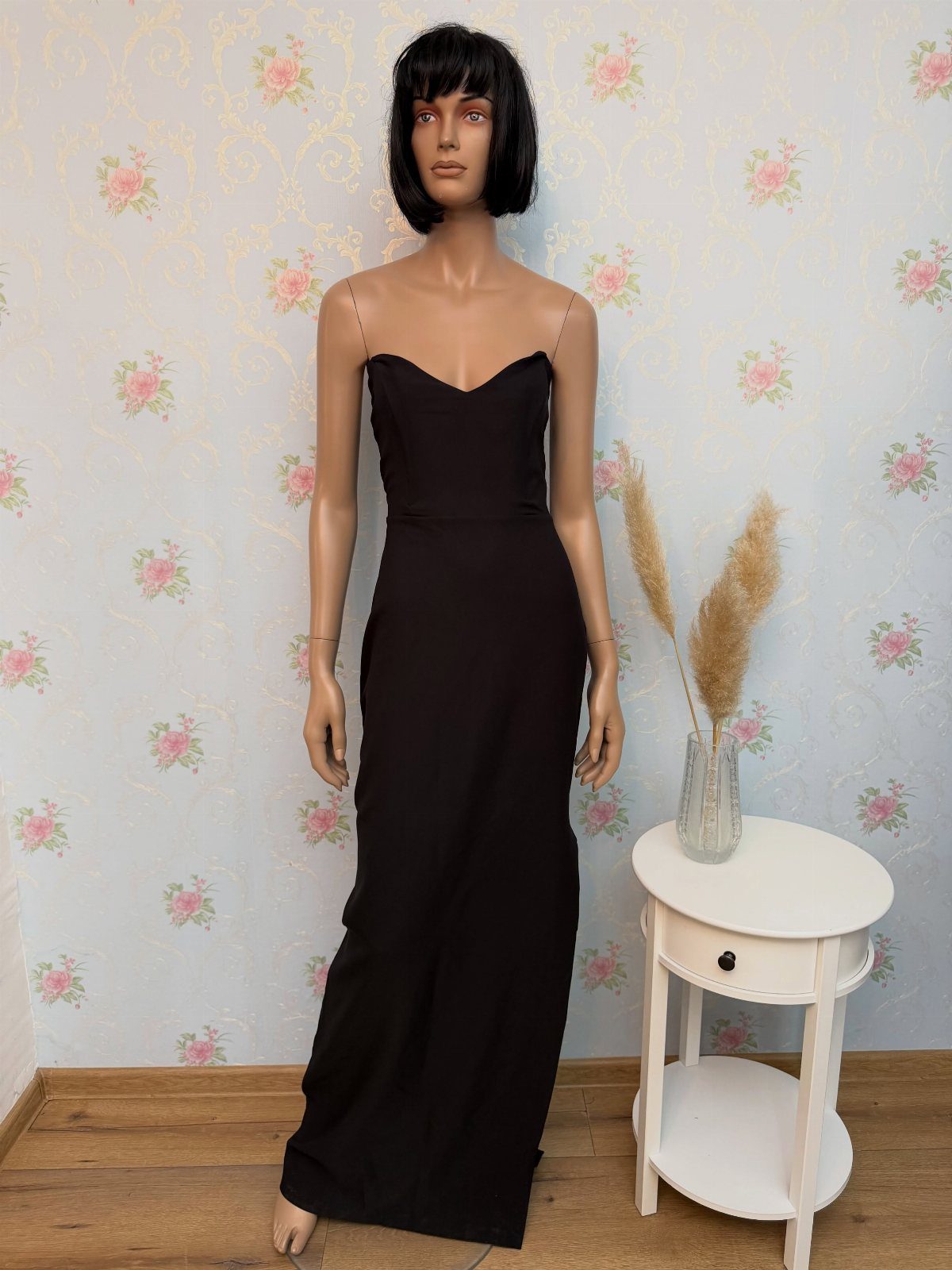 A black evening gown displayed on a mannequin against floral wallpaper.