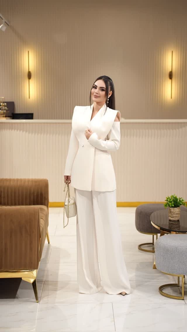 A woman posing in a white formal pantsuit and wide-leg trousers in a modern lounge.