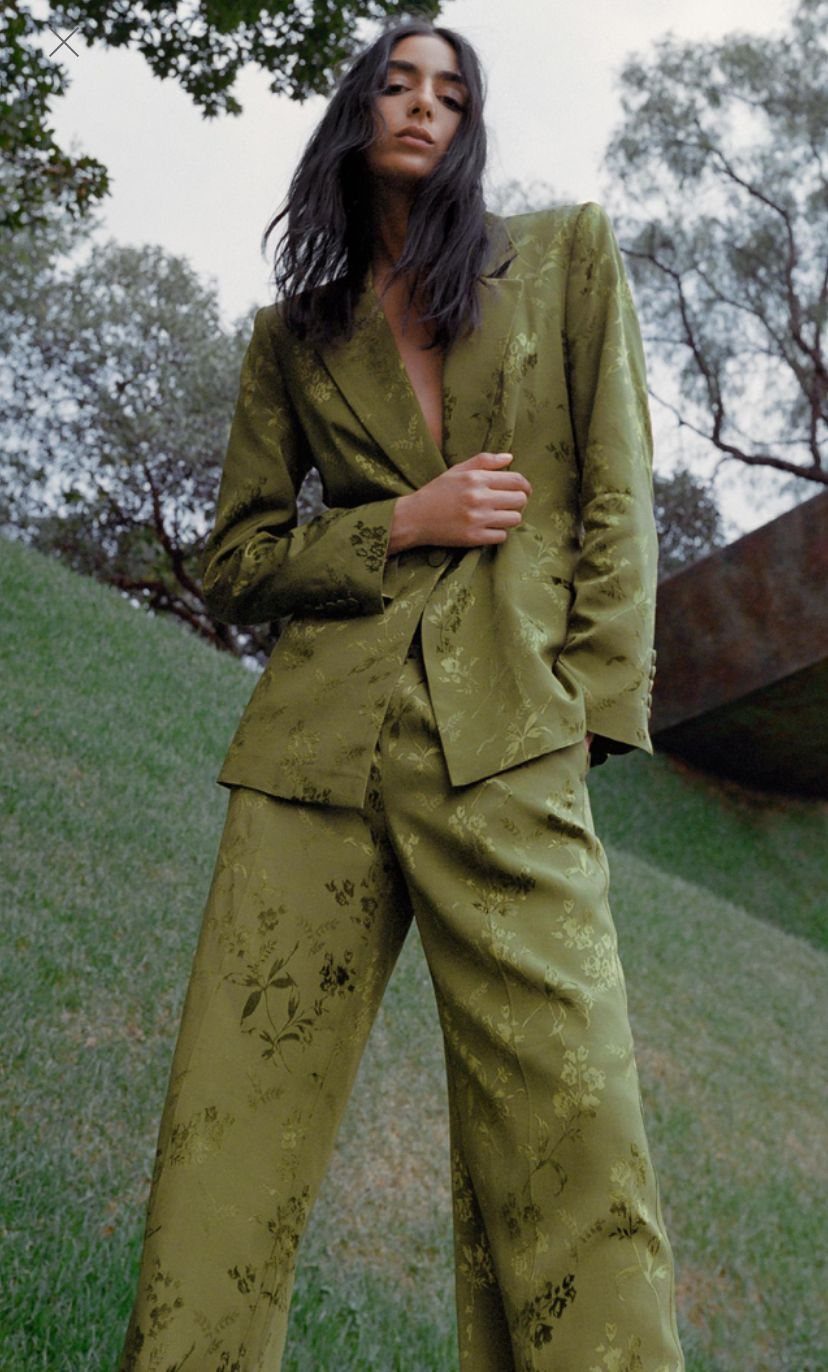 Woman wearing a tailored olive green floral jacquard pant suit in a grassy outdoor setting.