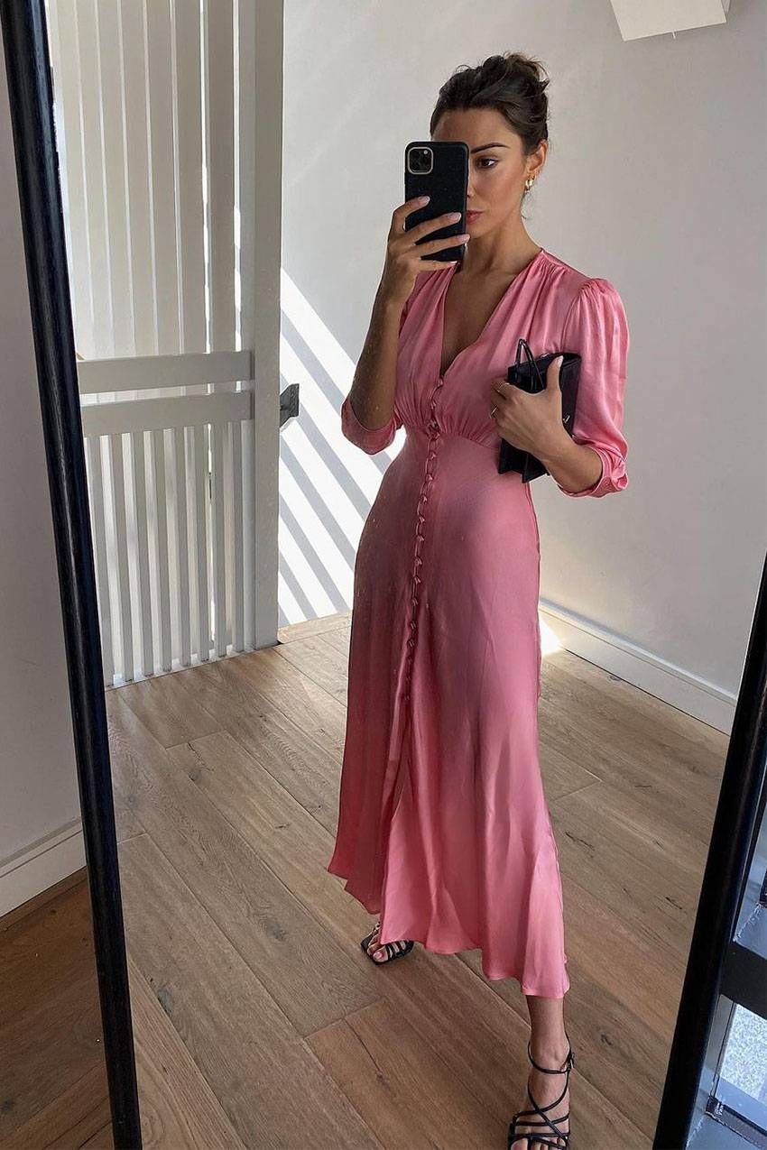 Woman in a long pink satin dress taking a mirror selfie in a room.