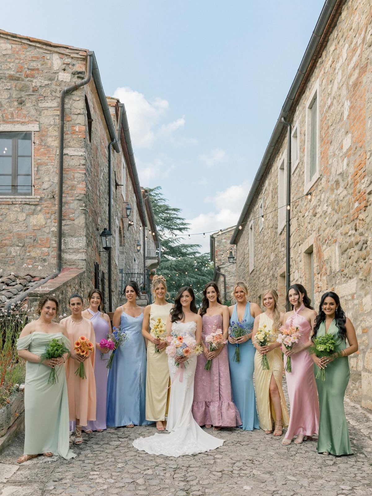 Bridesmaids in various pastel colored formal dresses standing in a stone alleyway holding bouquets.