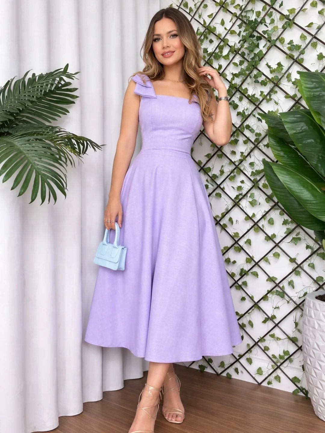 Woman in a lilac sleeveless midi dress posing with a small blue handbag against a leafy trellis.