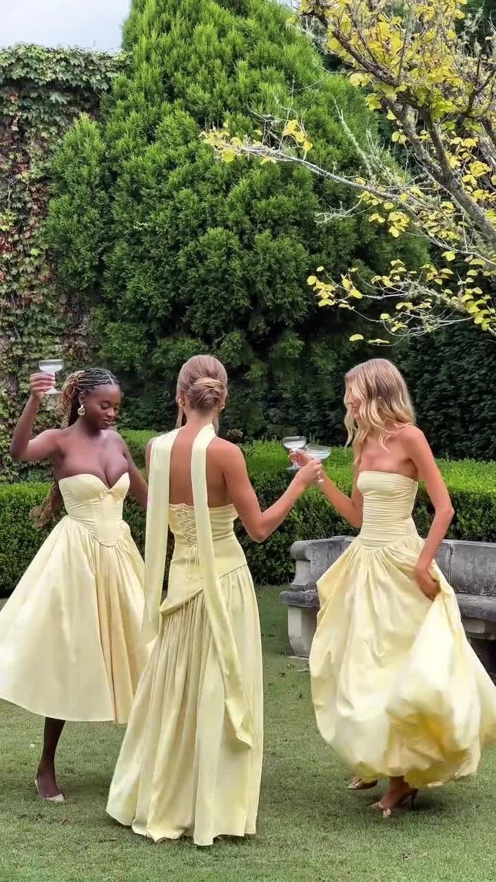 Three women in flowing pale yellow formal dresses enjoying cocktails in a lush green garden.