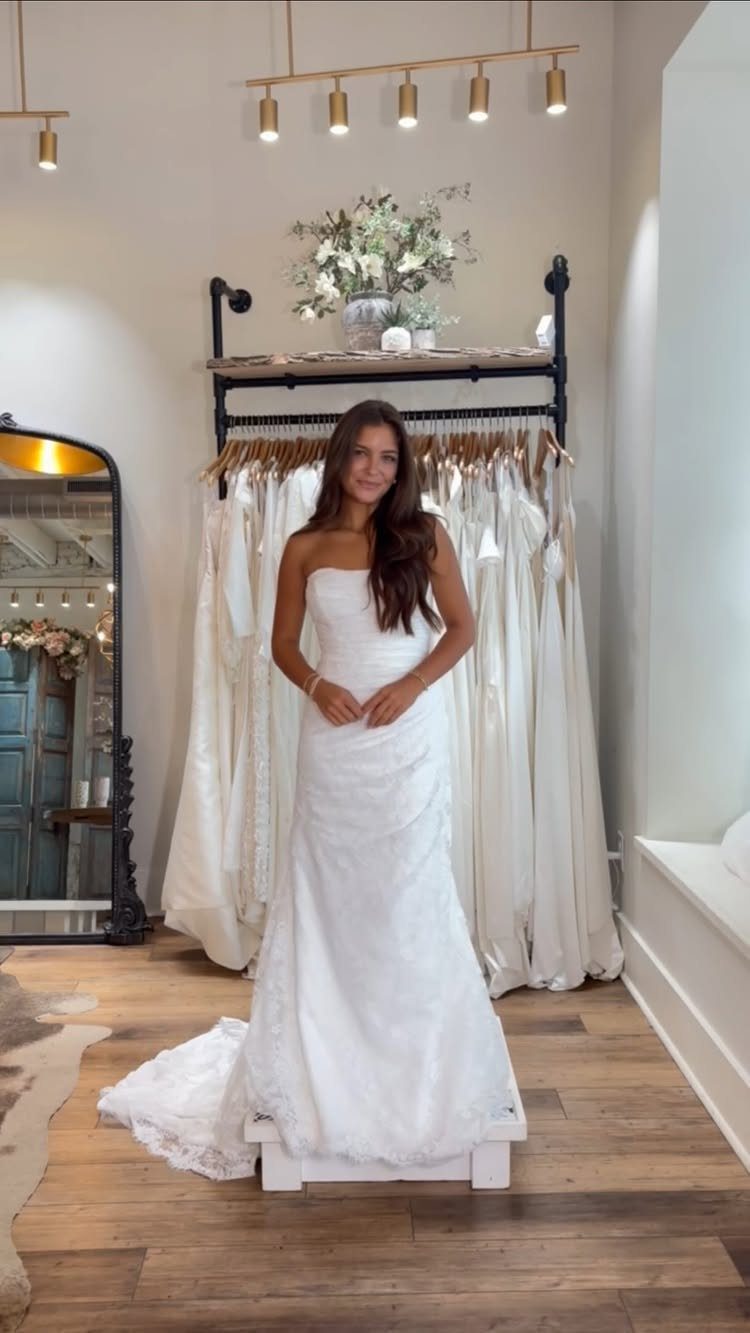 A woman posing in a white strapless lace wedding dress with a flared skirt.