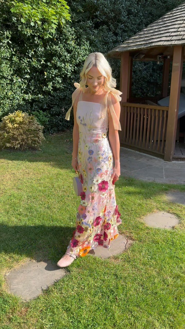 A blonde woman walking in a garden wearing a colorful floral maxi dress.