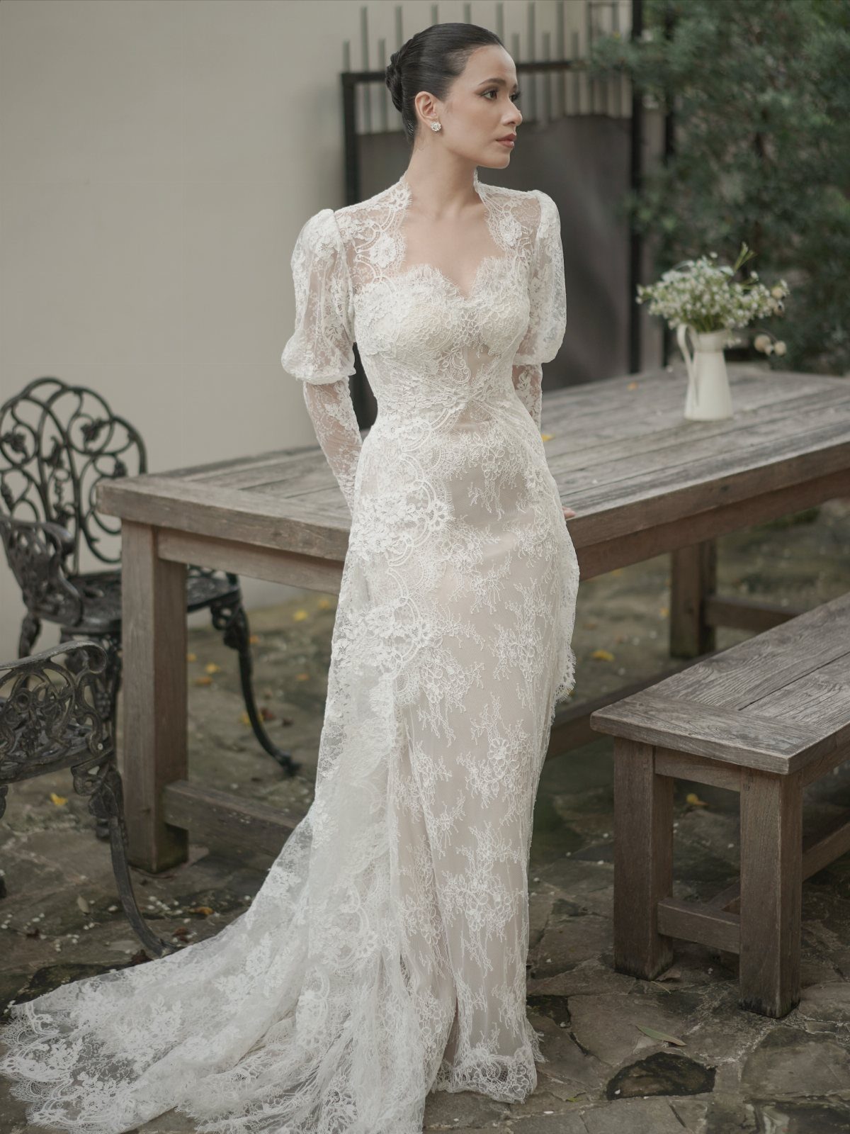 Woman in a white lace wedding dress standing by a wooden table in a garden setting.