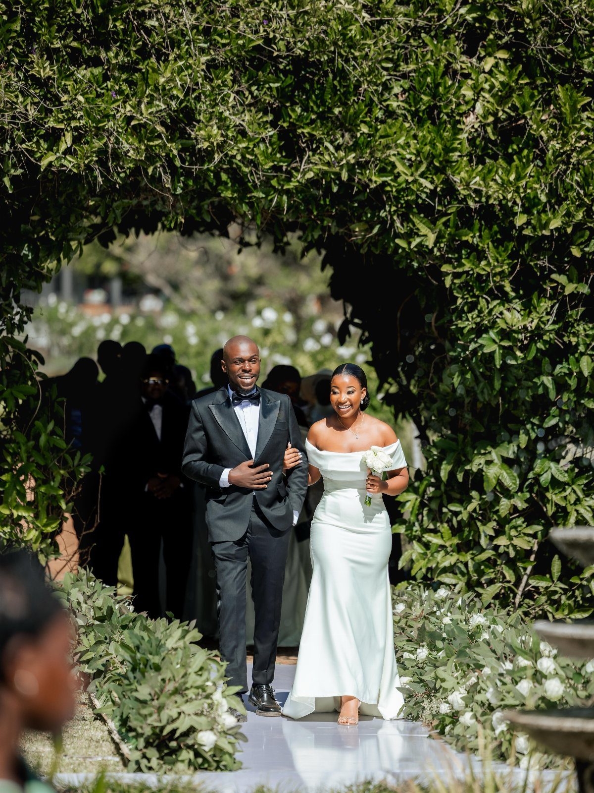 A bride in an off-the-shoulder white gown and a groom walking through a garden archway.