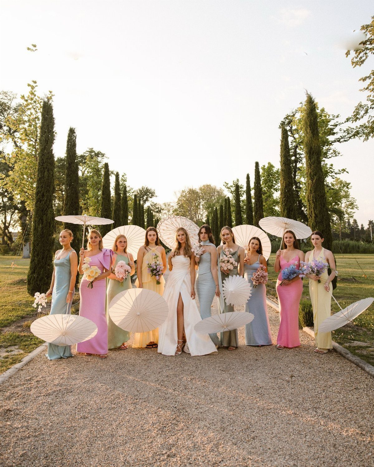 Bridesmaids in pastel gowns holding parasols and flowers on a gravel path lined with cypress trees.