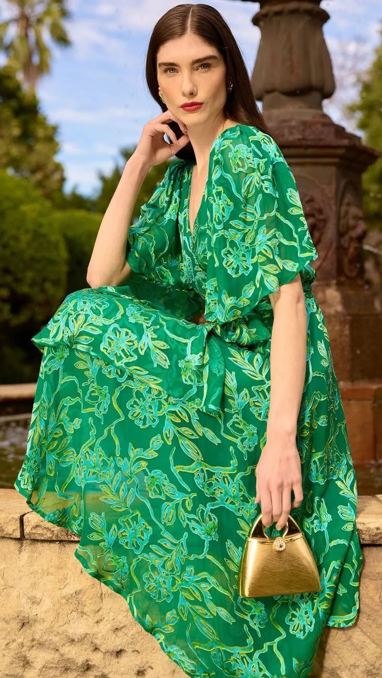 Woman in a green floral wrap dress sitting outdoors with a gold handbag.