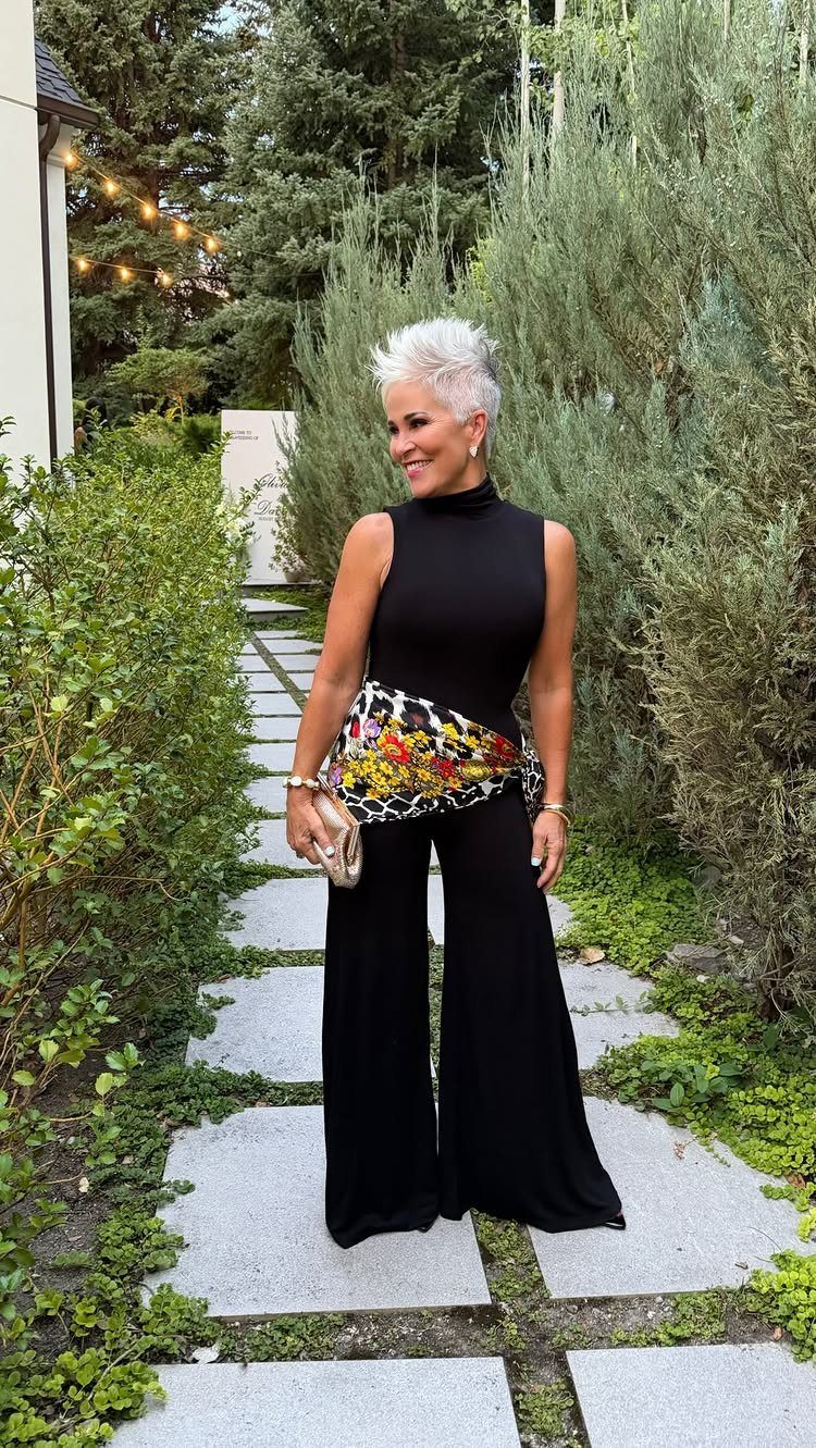 Woman in a black sleeveless jumpsuit with a colorful floral waist detail standing in a garden.