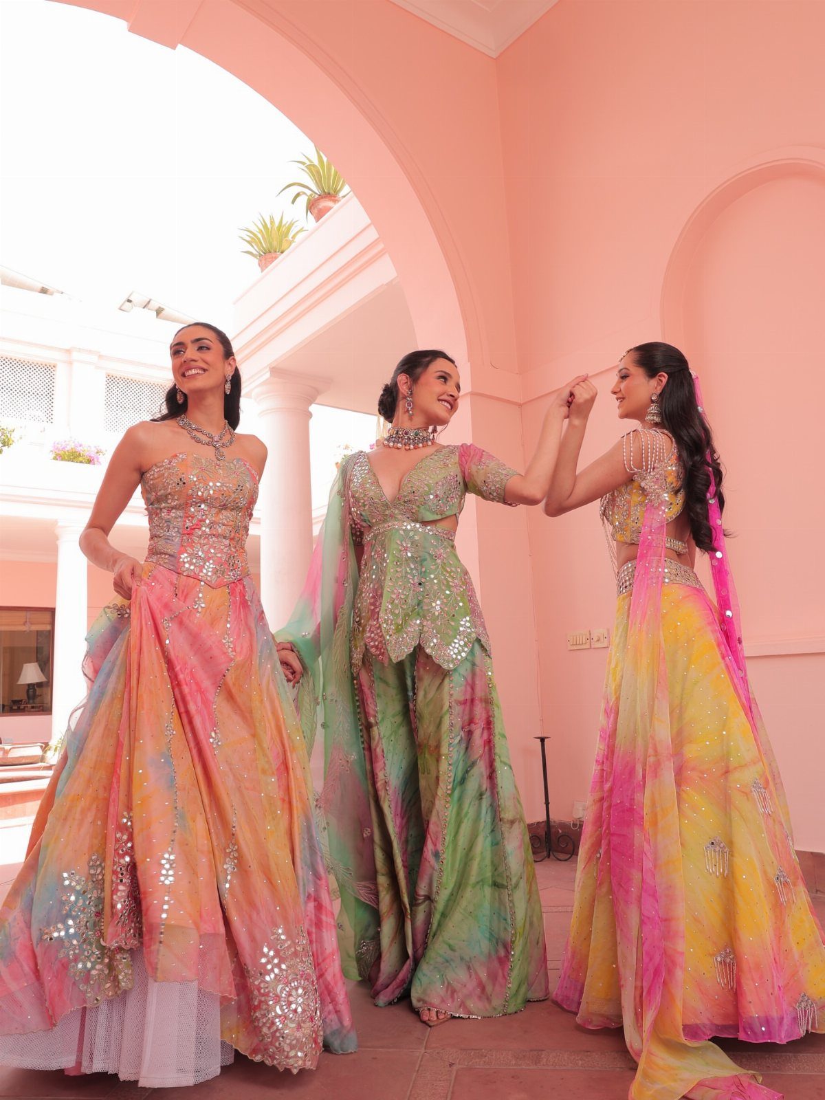 Three women wearing colorful, mirror-embellished Indian lehengas posing under a pink architectural archway.