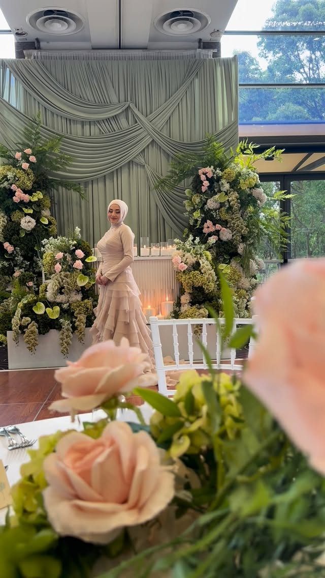 Woman in a tiered peach gown and white hijab standing amidst floral wedding decorations.