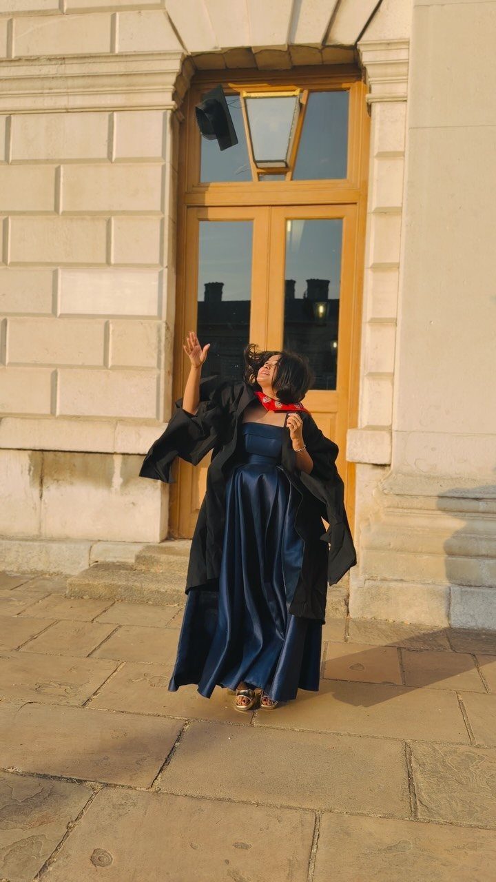 A graduate in a navy satin dress throws a cap into the air outside a building.