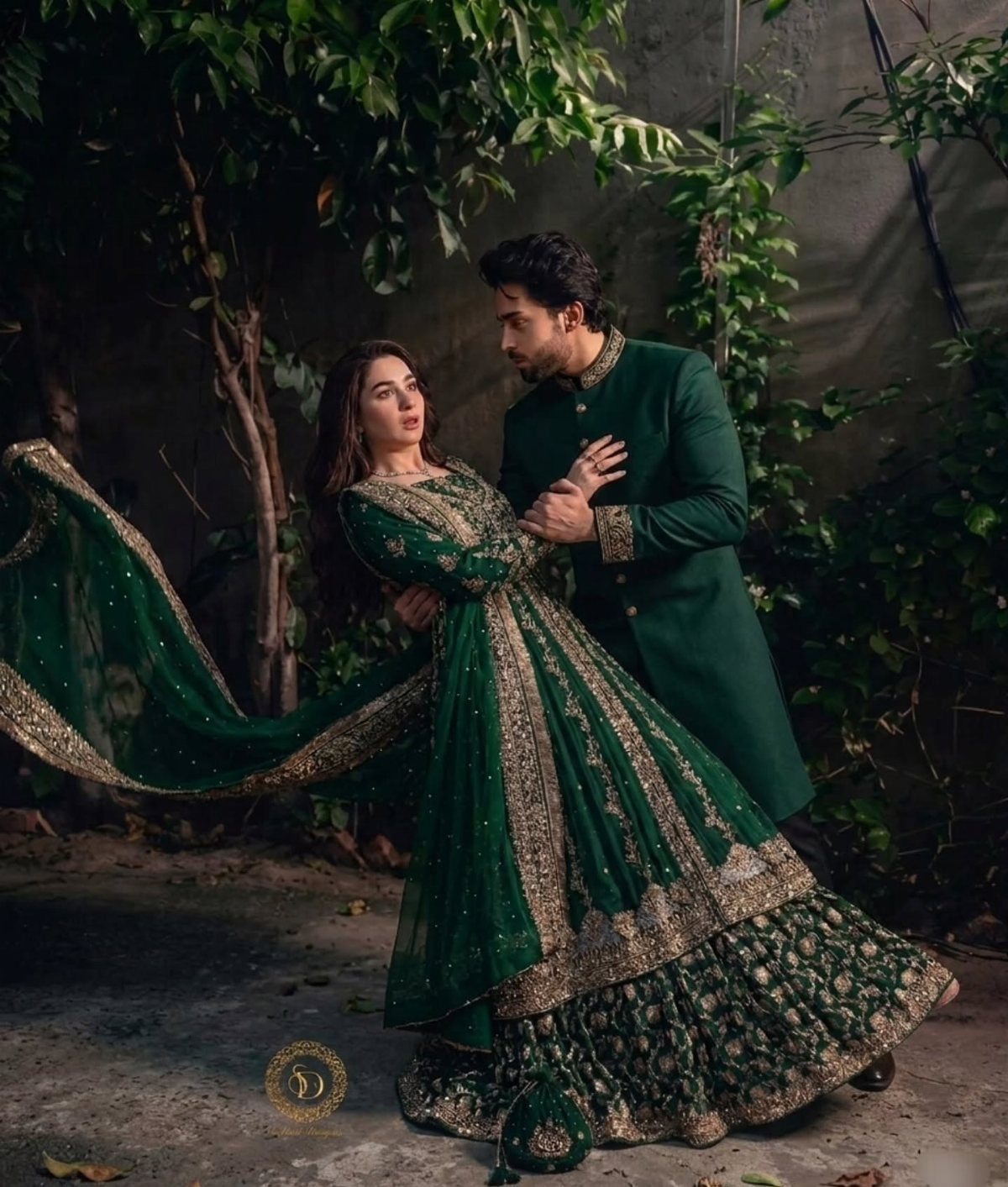 A man and woman posing in elegant, heavily embroidered emerald green formal South Asian attire amidst lush greenery.