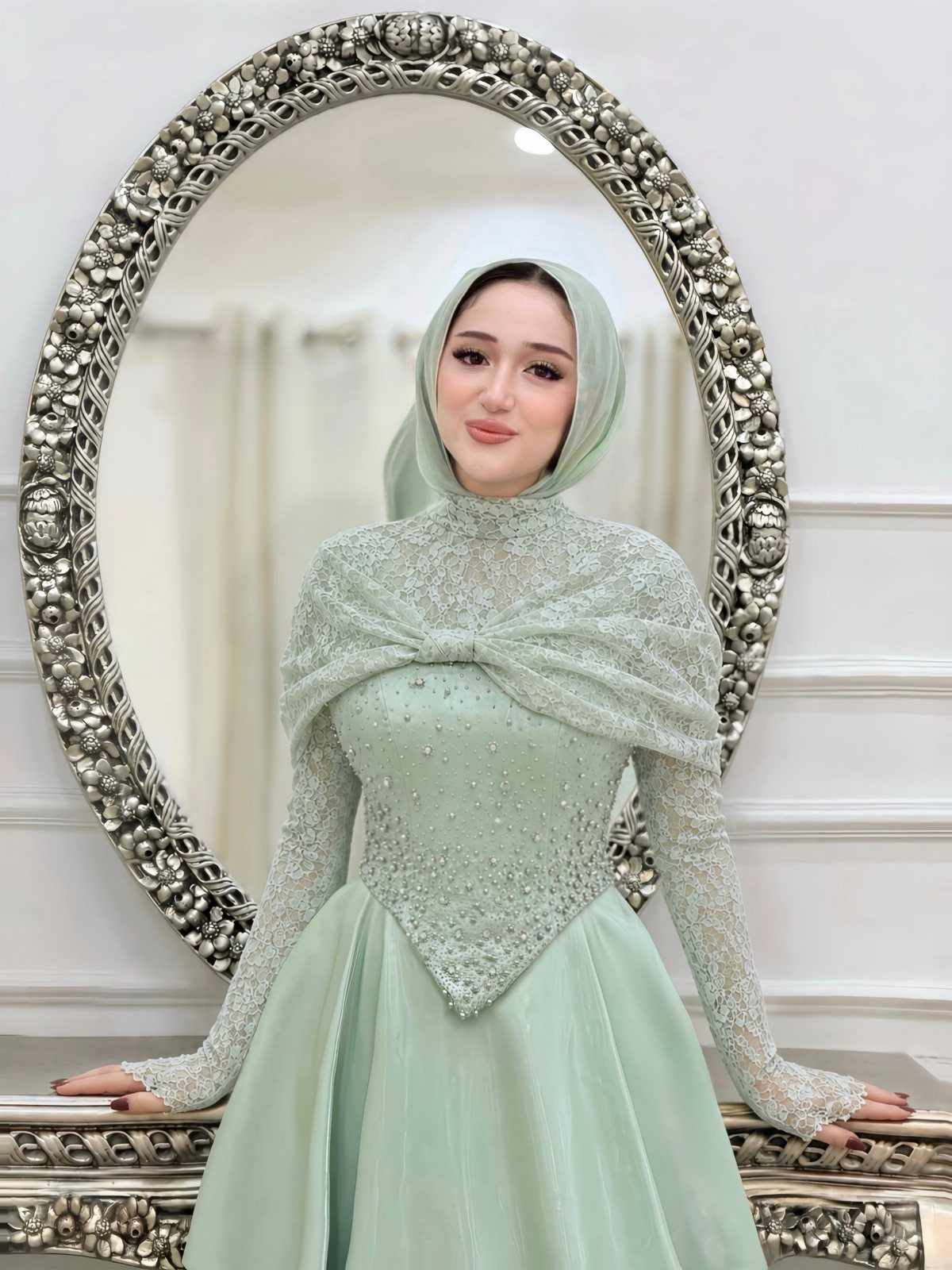 Woman in a mint green beaded formal gown and matching hijab posing by an ornate mirror.