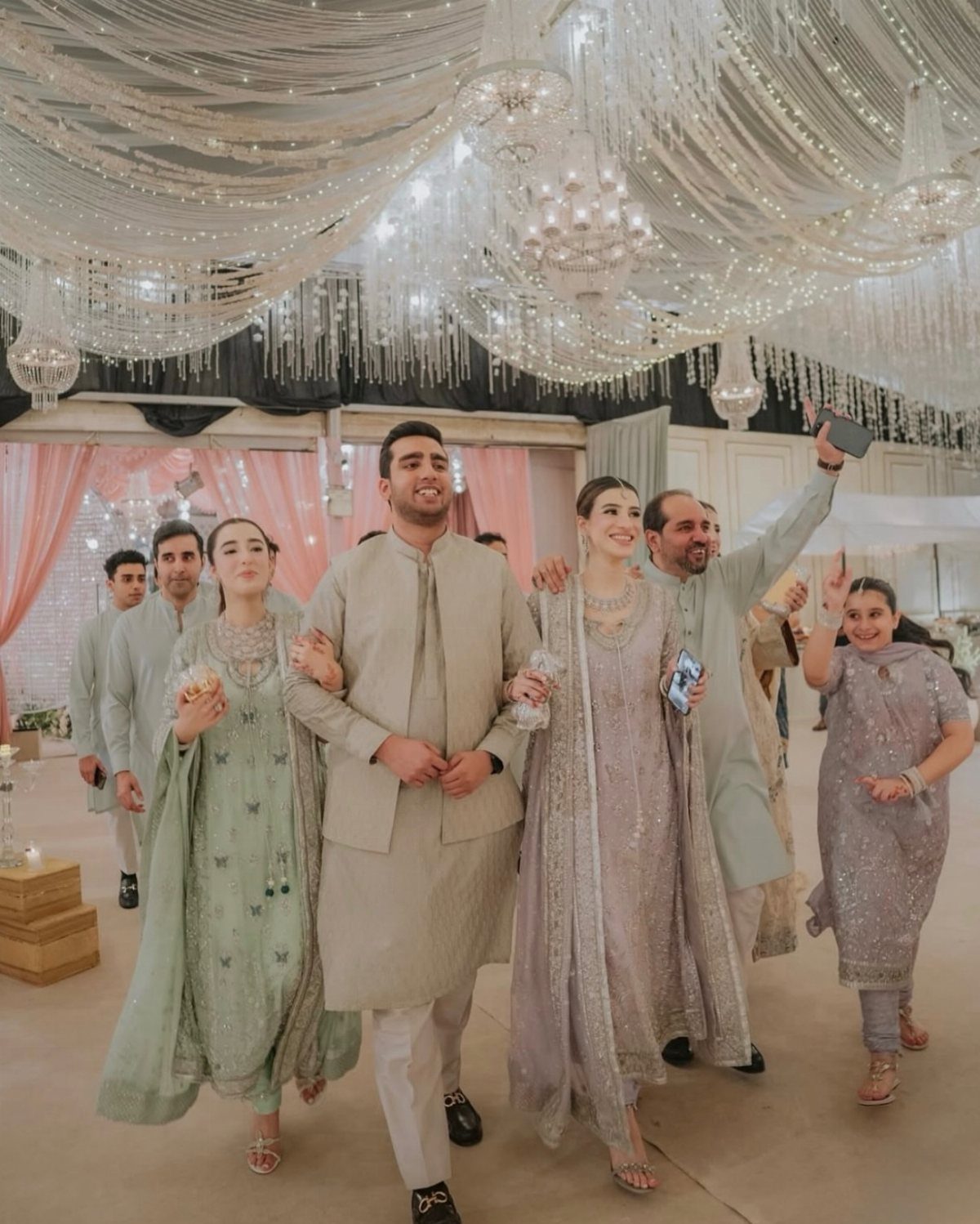 A woman in a mint green embroidered salwar kameez walking with a group in a decorated hall.