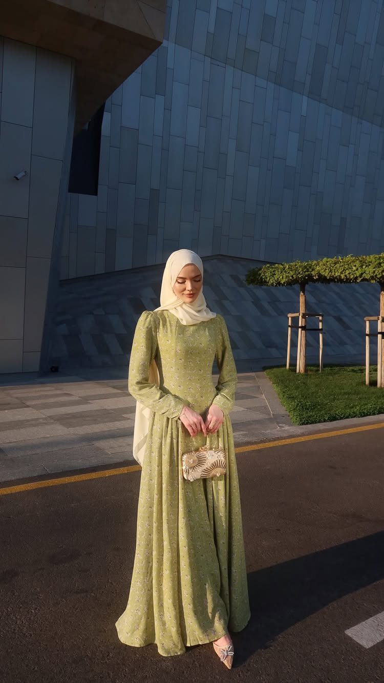 Woman in a long green dress and cream hijab holding a small clutch bag.