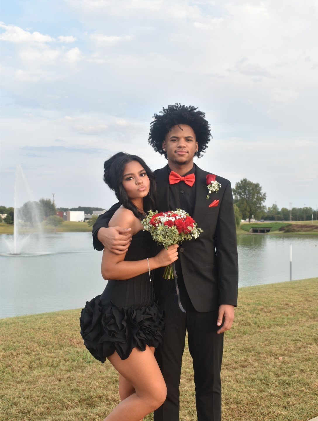 A young couple posing together outdoors near a lake, the woman wearing a black floral dress.