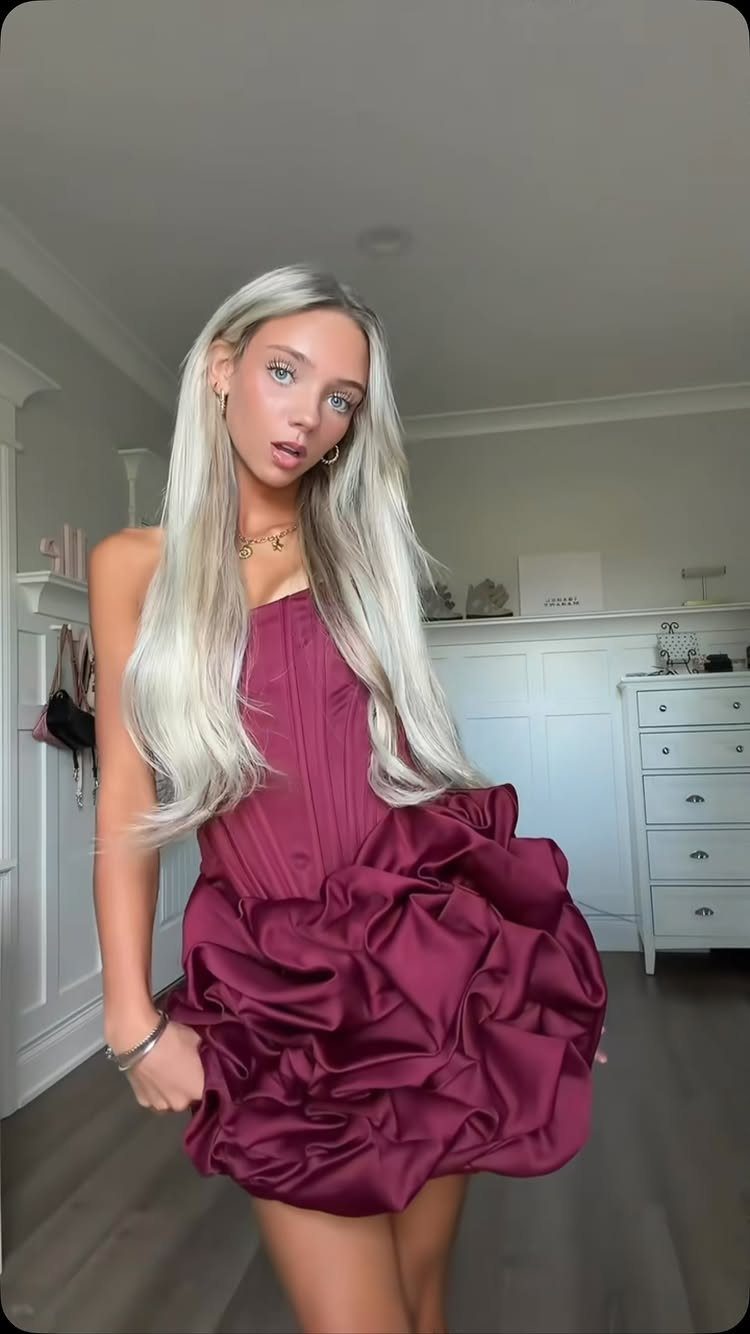 A young woman with long blonde hair posing in a dark magenta ruffled cocktail dress.