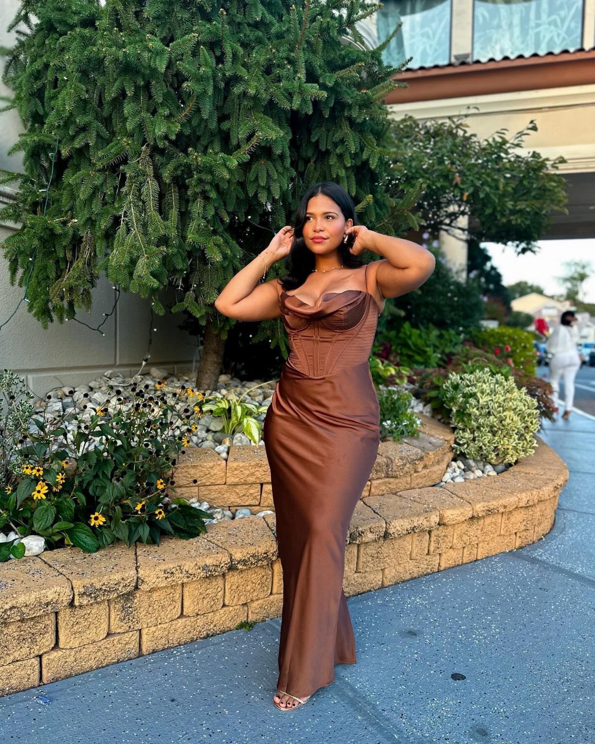 Woman in a brown satin corset maxi dress posing next to a stone planter and evergreen tree.